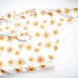 May include: White long-sleeved onesie with a yellow sun pattern.
