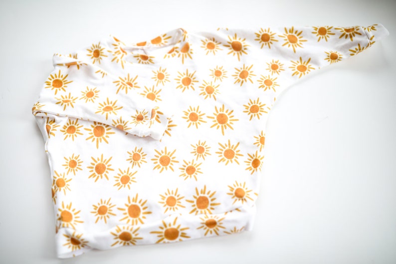 May include: White long-sleeved onesie with a yellow sun pattern.