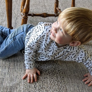 May include: A young child with blonde hair wearing a white shirt with black abstract patterns and blue jeans is lying on a beige carpet.