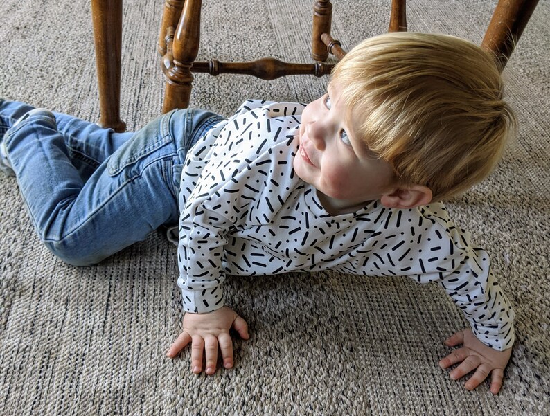 May include: A young child with blonde hair wearing a white shirt with black abstract patterns and blue jeans is lying on a beige carpet.