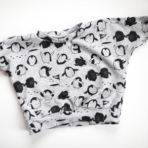 May include: A gray sweatshirt with a black penguin pattern. The penguins are in various poses, some are standing, some are swimming, and some are holding flags.