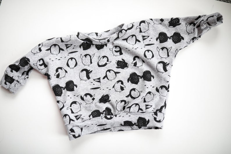 May include: A gray sweatshirt with a black penguin pattern. The penguins are in various poses, some are standing, some are swimming, and some are holding flags.