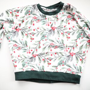May include: A white long-sleeved sweatshirt with a green and red holly berry print. The sweatshirt has a green ribbed neckline and cuffs.