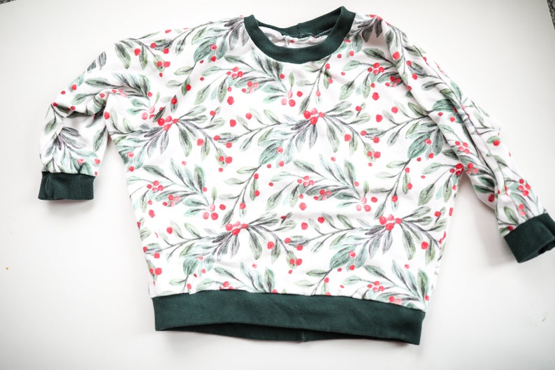 May include: A white long-sleeved sweatshirt with a green and red holly berry print. The sweatshirt has a green ribbed neckline and cuffs.