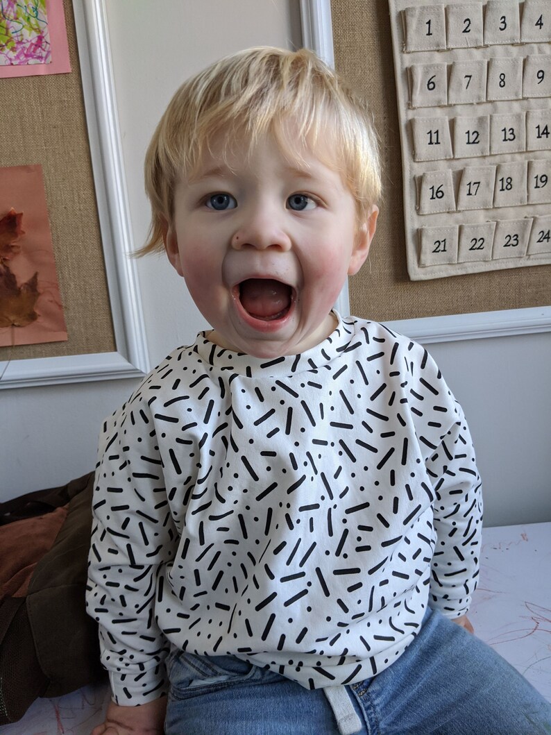 May include: A young child wearing a white long-sleeved shirt with a black and white confetti pattern. The child is wearing blue jeans and has blonde hair. The child is smiling and has their mouth open.