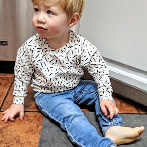 May include: A young child with red hair is sitting on the floor wearing a white long-sleeved shirt with a black and white confetti pattern and blue jeans.