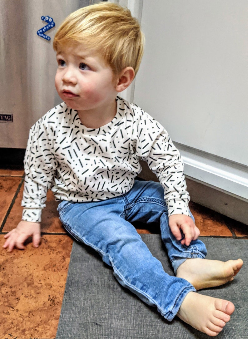 May include: A young child with red hair is sitting on the floor wearing a white long-sleeved shirt with a black and white confetti pattern and blue jeans.