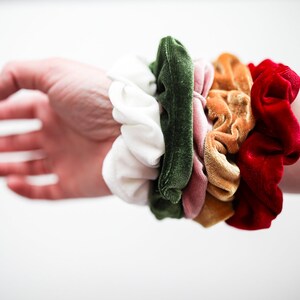 May include: A stack of colorful scrunchies in white, green, pink, orange, and red. The scrunchies are made of velvet and are arranged in a circle on a person's wrist.