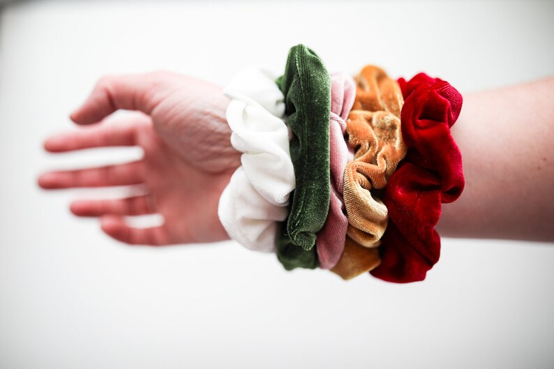 May include: A stack of colorful scrunchies in white, green, pink, orange, and red. The scrunchies are made of velvet and are arranged in a circle on a person's wrist.