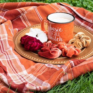 May include: A gold-colored tray with a white mug that says "basic fall witch" and several velvet scrunchies in shades of red, orange, and brown. The tray is on a plaid blanket.