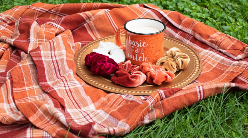 May include: A gold-colored tray with a white mug that says "basic fall witch" and several velvet scrunchies in shades of red, orange, and brown. The tray is on a plaid blanket.