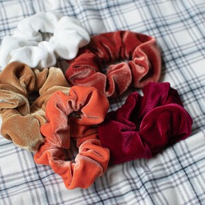 May include: A set of six colorful scrunchies in various shades of red, orange, brown, and white. The scrunchies are made of soft velvet and are perfect for holding hair in place.