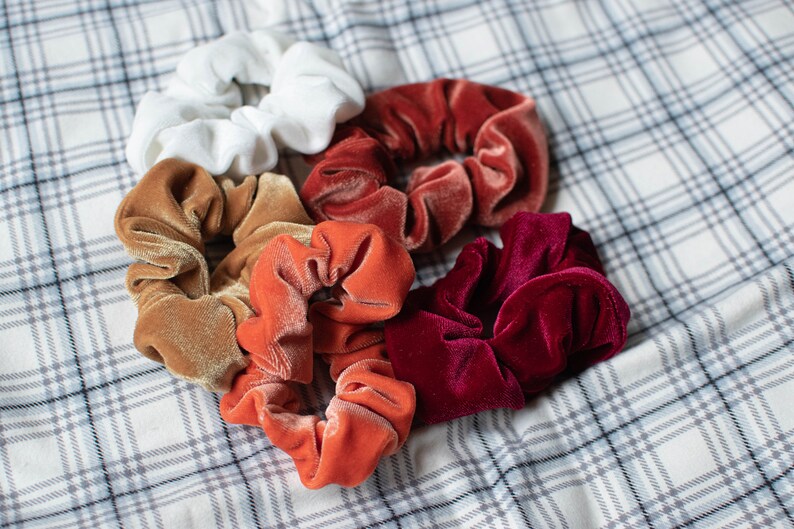 May include: A set of six colorful scrunchies in various shades of red, orange, brown, and white. The scrunchies are made of soft velvet and are perfect for holding hair in place.