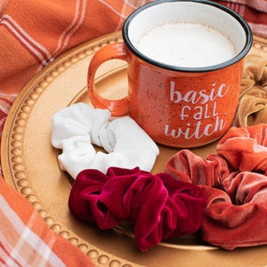 May include: A mug with the text "basic fall witch" on a gold plate with four velvet scrunchies in red, orange, white, and brown.