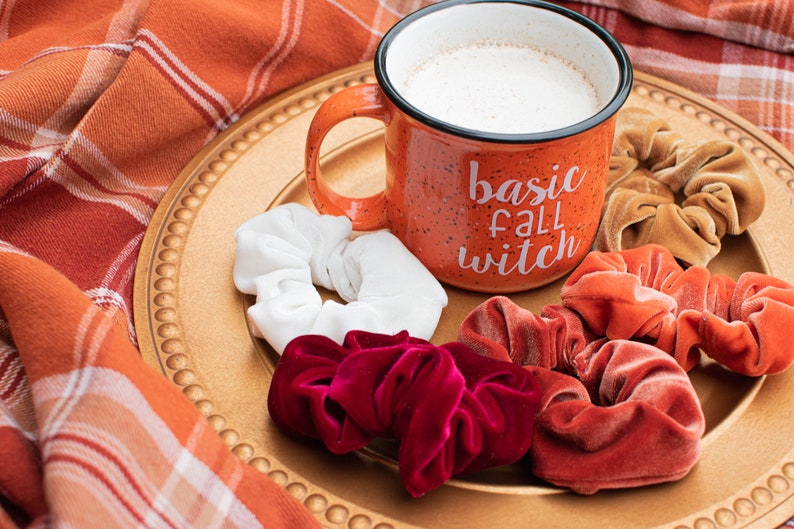 May include: A mug with the text "basic fall witch" on a gold plate with four velvet scrunchies in red, orange, white, and brown.