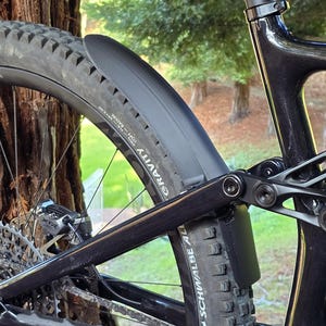 Puede incluir: Primer plano de la rueda trasera de una bicicleta de montaña negra con un neumático negro y un guardabarros negro. El neumático tiene la palabra "GRAVITY" impresa. La bicicleta está apoyada en un árbol.