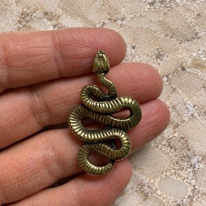 May include: A brass-toned metal pendant shaped like a coiled snake. The snake is depicted in a winding, S-shaped position with a textured surface that mimics scales. The metal has a slightly weathered, antique finish with dark highlights in the crevices.