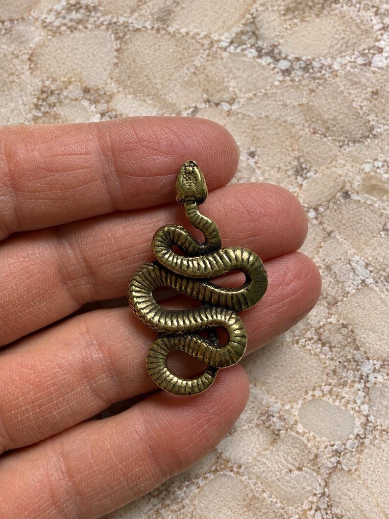 May include: A brass-toned metal pendant shaped like a coiled snake. The snake is depicted in a winding, S-shaped position with a textured surface that mimics scales. The metal has a slightly weathered, antique finish with dark highlights in the crevices.
