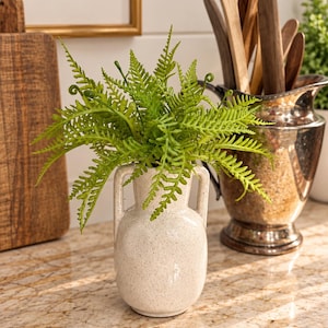 May include: A small, cream-colored ceramic vase with two side handles, featuring a speckled texture. The vase contains a vibrant green artificial fern plant with detailed, feathery fronds that extend outward from the center opening.