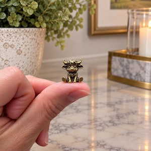 May include: A miniature brass-colored figurine of a Highland cow with shaggy hair, curved horns, and a seated posture. The small metal sculpture features detailed facial expressions and textured fur, captured in a compact, three-dimensional form.