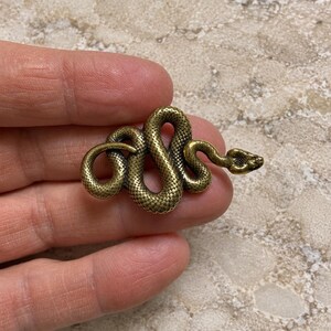 May include: A small, antique-style brass-toned metal snake figurine with a coiled body and textured scales. The snake is depicted in a winding, serpentine position with its head slightly raised, showcasing a detailed, realistic surface finish.