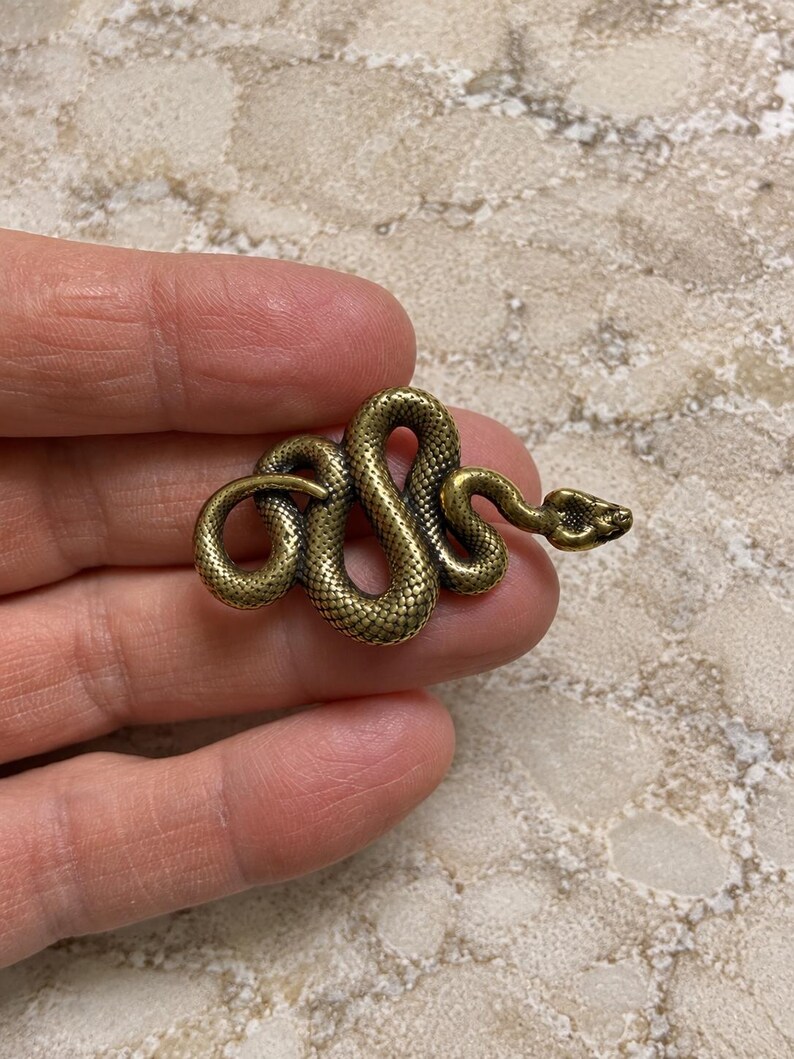 May include: A small, antique-style brass-toned metal snake figurine with a coiled body and textured scales. The snake is depicted in a winding, serpentine position with its head slightly raised, showcasing a detailed, realistic surface finish.
