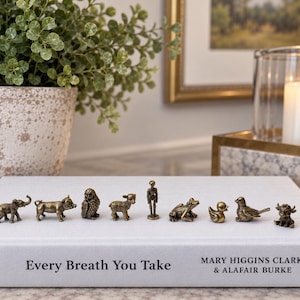 May include: A collection of nine small bronze-toned metal animal and human figurines arranged in a row on top of a white hardcover book titled Every Breath You Take by Mary Higgins Clark and Alafair Burke. The figures include an elephant, pig, owl, lamb, person, frog, duck, bird, and bull.