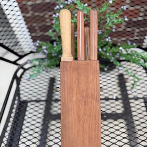 May include: A rectangular wooden knife block containing three knives with wooden handles. The block is made of a medium-brown wood, while the knife handles vary in shade from light tan to a deeper reddish-brown, each featuring a thin metal bolster.