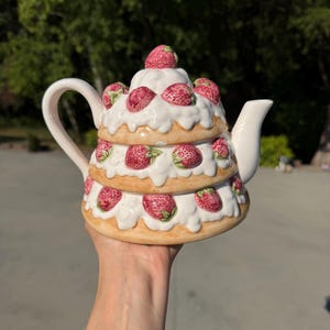 Può includere: Una teiera in ceramica a forma di torta a tre piani con glassa bianca e fragole rosse. La teiera presenta un manico curvo, un beccuccio e un coperchio sormontato da una singola fragola. Il design include diverse fragole scolpite.