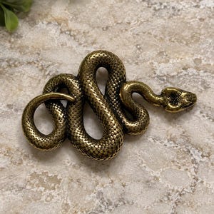 May include: A metallic gold-toned snake figurine with a textured, scaled surface. The snake is coiled into an S-shape with its head raised and pointed toward the right side, showcasing detailed eyes and a defined snout in a realistic posture.