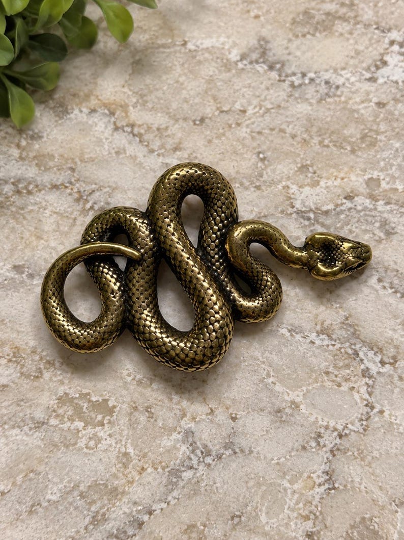 May include: A metallic gold-toned snake figurine with a textured, scaled surface. The snake is coiled into an S-shape with its head raised and pointed toward the right side, showcasing detailed eyes and a defined snout in a realistic posture.