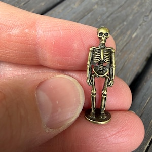 May include: A miniature antique brass-colored metal skeleton figurine standing on a small circular base. The figure features detailed molded bones, including a skull, rib cage, and limbs, with a weathered metallic finish throughout the entire structure.