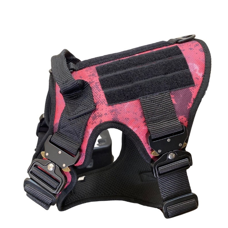 May include: A pink and black tactical dog harness with adjustable straps and a buckle closure. The harness has a black patch on the back with a hook and loop fastener.