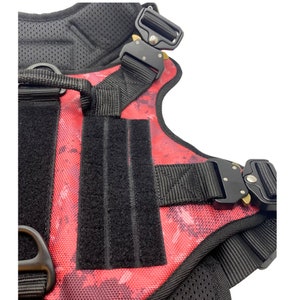 May include: A red and black dog harness with adjustable straps and a buckle closure. The harness has a camouflage pattern and a black velcro patch.