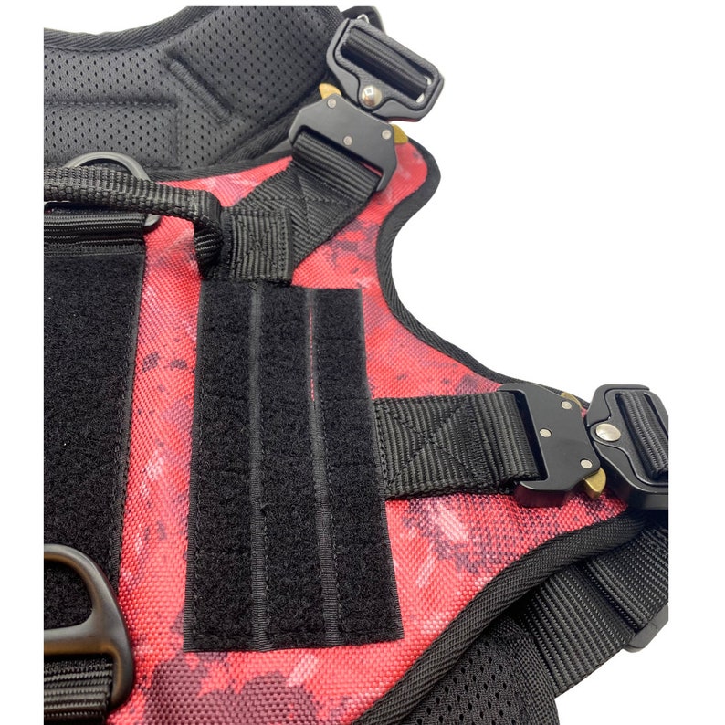 May include: A red and black dog harness with adjustable straps and a buckle closure. The harness has a camouflage pattern and a black velcro patch.