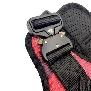 May include: Close-up of a black adjustable buckle with a red and black patterned strap. The buckle has a metal release lever and a small square logo.
