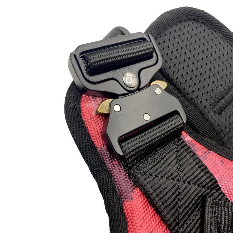 May include: Close-up of a black adjustable buckle with a red and black patterned strap. The buckle has a metal release lever and a small square logo.