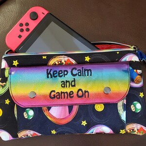 Peut inclure: Une pochette de jeu noire et arc-en-ciel avec une console de jeu rose et noire. La pochette a un rabat arc-en-ciel avec le texte "Keep Calm and Game On". La pochette est décorée de graphismes sur le thème des jeux vidéo.