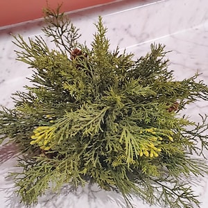 May include: A green artificial cedar sprig with yellow flower accents. The sprig is on a white marble surface.