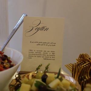Könnte beinhalten: Eine Tischdekoration mit einer weißen Karte mit Kalligraphie und Text, neben einer Schüssel mit Salat und einem Teller mit Oliven und Käse. Goldfarbene Gabeln sind im Hintergrund zu sehen.