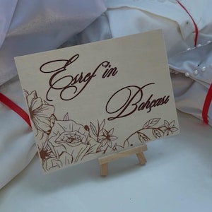 May include: A rectangular light-colored wooden sign resting on a small wooden easel. The sign features dark brown engraved text that reads Esref'in Bahçesi, accompanied by a detailed floral illustration of roses and leaves at the bottom left corner.