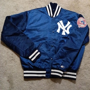 May include: A navy blue satin jacket with white detailing, displaying the New York Yankees logo and a team patch. The jacket has a button-up front, striped cuffs, and a striped hem. This vintage sports jacket is a classic piece of apparel.