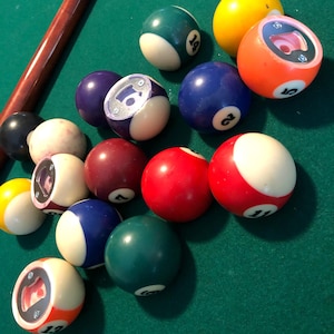 Puede incluir: Una colección de bolas de billar con abridores de botellas sobre una mesa de billar de fieltro verde. Las bolas son de varios colores, incluyendo rojo, amarillo, azul y blanco. Los números 1, 5, 6, 7, 11 y 14 son visibles en las bolas.