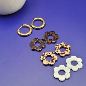 Puede incluir: Un conjunto de seis pendientes, que incluye dos pendientes de aro dorados, dos pendientes florales marrones y negros y dos pendientes florales blancos.