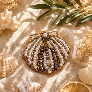 Può includere: Una spilla a forma di conchiglia realizzata con file di perle bianche e perline color bronzo metallizzato. Il design presenta una linea verticale centrale di cristalli trasparenti e una singola perla bianca sulla parte superiore.