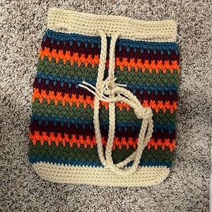 May include: A crocheted bag with a striped pattern in shades of green, orange, burgundy, and blue. The bag has a cream-colored top and a long cream-colored strap.