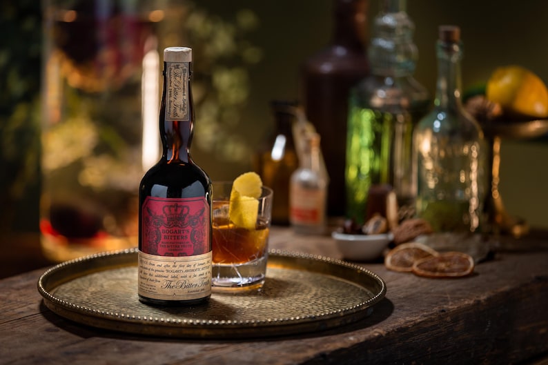 May include: A dark brown bottle of "Sogarth Bitters" with a red label and gold accents sits on a gold tray. A cocktail with a lemon peel garnish is next to the bottle. Other bottles and garnishes are in the background.