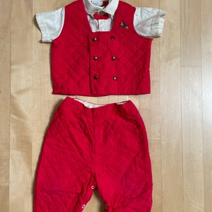 May include: A red quilted vest and matching pants set for a baby or toddler. The vest has a double breasted closure with gold buttons and a white shirt with a bow tie underneath. The pants have a snap closure at the bottom.