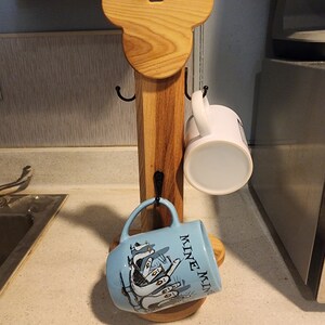 May include: Wooden mug rack shaped like Mickey Mouse head with two hooks on each side and a round base. Two mugs are hanging on the hooks, one is white and the other is blue with a design of penguins and the text "Mine Mine Mine".