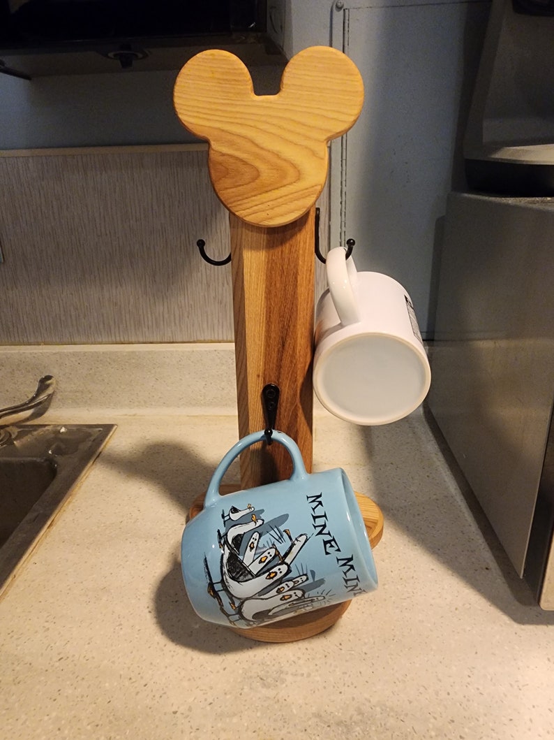 May include: Wooden mug rack shaped like Mickey Mouse head with two hooks on each side and a round base. Two mugs are hanging on the hooks, one is white and the other is blue with a design of penguins and the text "Mine Mine Mine".